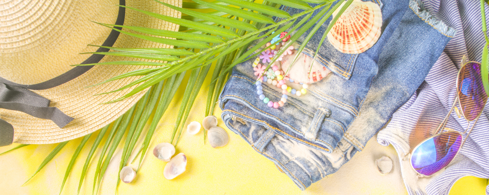 Kurze Jeans, Accessoires und ein Strohhut liegen mit einem Palmenblatt im Sand