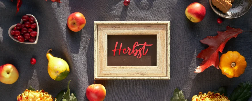 Ein Holzschild mit der Aufschrift Herbst in roter Schreibschrift auf dunklem Hintergrund. Das Schild liegt auf einem dunklen Tisch, umgeben von Früchten und herbstlicher Deko.